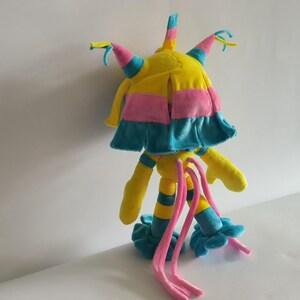 K&ouml;nnte beinhalten: Ein Pl&uuml;schtier in den Farben Gelb, Pink und Blau. Die Figur hat einen zentralen Kopf mit mehreren spitzen Forts&auml;tzen, ein schichtartiges Element, lange, d&uuml;nne Gliedma&szlig;en mit Streifenmuster und zwei lange, herabh&auml;ngende pinke Anh&auml;ngsel.