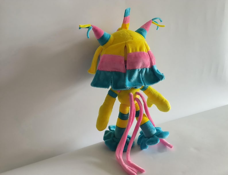 K&ouml;nnte beinhalten: Ein Pl&uuml;schtier in den Farben Gelb, Pink und Blau. Die Figur hat einen zentralen Kopf mit mehreren spitzen Forts&auml;tzen, ein schichtartiges Element, lange, d&uuml;nne Gliedma&szlig;en mit Streifenmuster und zwei lange, herabh&auml;ngende pinke Anh&auml;ngsel.