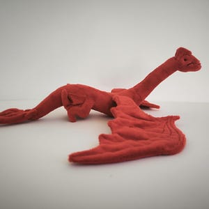 Pode incluir: Um brinquedo de pelúcia de dragão vermelho com um pescoço longo, uma cabeça pequena e asas largas estendidas. A criatura apresenta uma superfície de tecido texturizado, um corpo esguio e uma cauda que afunila, tudo em uma cor vermelha sólida.
