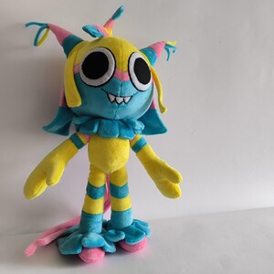K&ouml;nnte beinhalten: Ein Pl&uuml;schtier in Form einer gelben, blauen und rosafarbenen Kreatur mit gro&szlig;en schwarzen Augen, einem breiten Grinsen mit sichtbaren Z&auml;hnen und einem R&uuml;schenkragen. Die Figur hat lange, d&uuml;nne Gliedma&szlig;en mit gelb-blauen Streifen und einen langen rosa Schwanz.