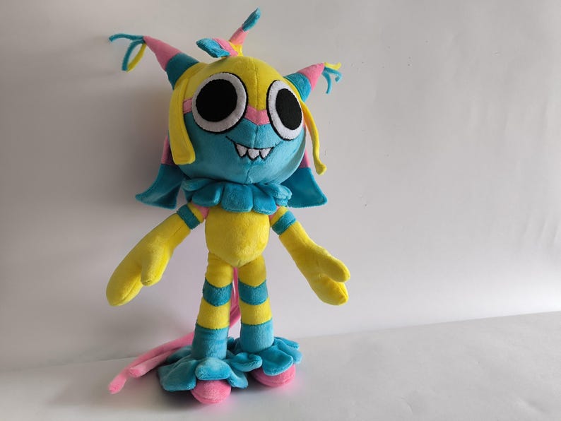 K&ouml;nnte beinhalten: Ein Pl&uuml;schtier in Form einer gelben, blauen und rosafarbenen Kreatur mit gro&szlig;en schwarzen Augen, einem breiten Grinsen mit sichtbaren Z&auml;hnen und einem R&uuml;schenkragen. Die Figur hat lange, d&uuml;nne Gliedma&szlig;en mit gelb-blauen Streifen und einen langen rosa Schwanz.