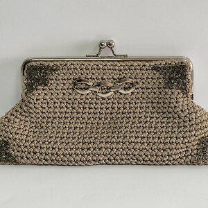 May include: A beige crocheted clutch with a silver clasp and decorative metal corner accents. The clutch features a silver chain detail on the front and a kiss-lock closure. This handmade bag is a stylish accessory.