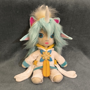 May include: A plush doll with a unicorn horn, blue hair, and large blue eyes. The doll wears a white outfit with yellow and blue accents, including a star-shaped emblem. The doll has pink ears and a fluffy, light-colored hairpiece.