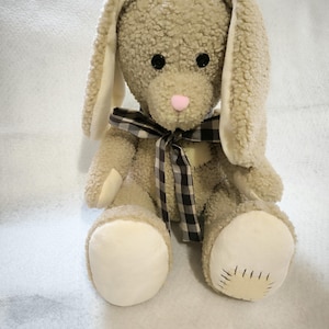 Puede incluir: Un conejo de peluche beige con orejas largas y caídas y una nariz rosa. El conejo tiene un cuerpo texturizado y difuso y detalles blancos en las patas y el interior de las orejas. Una cinta a cuadros en blanco y negro está atada alrededor del cuello.