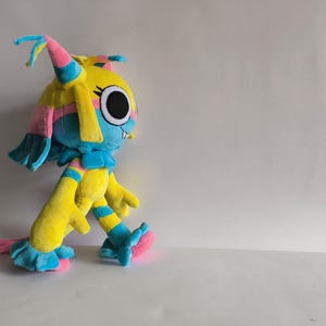 K&ouml;nnte beinhalten: Ein Pl&uuml;schtier mit einem leuchtend gelben Kopf und K&ouml;rper, verziert mit lebhaften blauen und rosafarbenen Akzenten. Die Figur hat gro&szlig;e, runde schwarze Augen, einen kleinen wei&szlig;en Zahn und lange, flie&szlig;ende haar&auml;hnliche Forts&auml;tze.