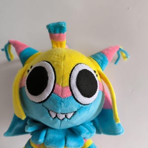 K&ouml;nnte beinhalten: Ein Pl&uuml;schtier mit einem leuchtend gelben Kopf, gro&szlig;en schwarzen runden Augen und einem breiten Grinsen mit wei&szlig;en Z&auml;hnen. Die Figur hat blaue und rosa Akzente, einschlie&szlig;lich spitzer Ohren mit herabh&auml;ngenden gelben Str&auml;hnen.