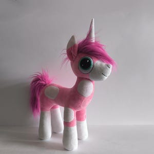 May include: A pink plush unicorn with white patches and legs. It features a pink mane and tail, a white horn, and large eyes. The toy has a friendly, cartoon-like design, suitable for children.