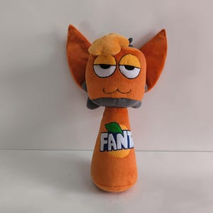 May include: Orange plush toy shaped like a cartoon character with large orange ears and a small hat. The character has a gray neck and a body shaped like a soda bottle with the word "FANTA" printed on it.