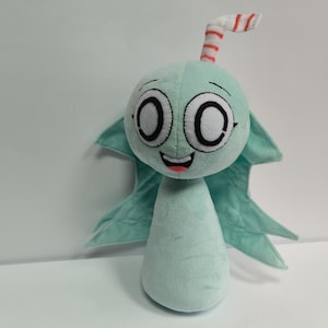 Puede incluir: Un peluche azul claro con forma de personaje de dibujos animados, con ojos grandes y redondos y una boca sonriente. Tiene pequeñas alas y una antena a rayas. Juguete infantil.