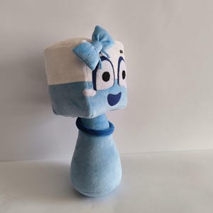 May include: A soft toy resembling a cartoon character. The head is a white and blue cube with large eyes, a blue bow, and a smiling mouth. The body is a light blue teardrop shape with a blue ring around the neck.