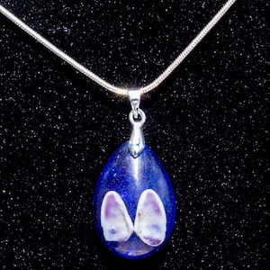 May include: A teardrop-shaped pendant necklace with a deep blue resin base. Two white and purple seashell-like shapes are embedded within the resin. The pendant hangs from a silver chain, set against a black background.