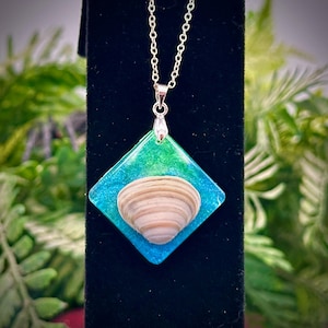 May include: A silver necklace featuring a pendant with a seashell encased in a square of blue and green resin. The pendant is suspended from a delicate silver chain, creating a coastal-themed accessory.
