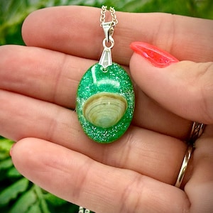 May include: Oval-shaped pendant necklace with a silver chain. The pendant features a green, glittery resin base with a seashell embedded in the center. The seashell is light brown and has a textured surface. The necklace is held in a hand.