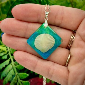 May include: A silver necklace featuring a square pendant with a seashell encased in resin. The pendant has a gradient of green and blue colors. The necklace is held in a hand with a green leafy background.