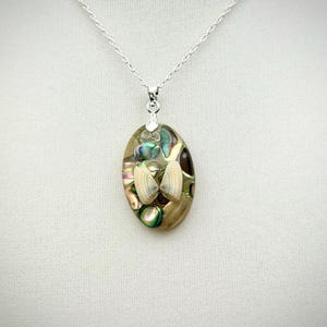May include: Oval pendant necklace with a silver chain. The pendant features a mosaic of iridescent abalone shell pieces and small seashells. The pendant is approximately 1.5 inches long and 1 inch wide.