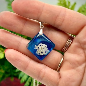 May include: A blue and white pendant necklace with a square, resin-filled charm. The charm features a white, coral-like design set against a deep blue background. The necklace has a silver-colored chain and clasp.