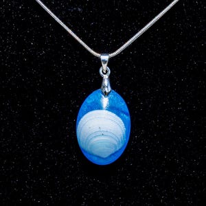 May include: Oval pendant necklace featuring a white seashell encased in blue resin. The pendant hangs from a delicate silver chain. The background is a dark, textured surface, highlighting the pendant's vibrant colors and ocean theme.