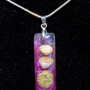 May include: A rectangular pendant necklace with three seashells encased in a clear, shimmering, pink resin. The pendant is suspended from a silver chain. The background is black.
