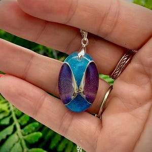 May include: Oval pendant necklace with a silver chain. The pendant features a vibrant blue background with a central design of purple and white elements. The necklace is held in a hand with red nail polish.