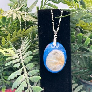 May include: A silver chain necklace with an oval pendant. The pendant features a blue background with a seashell design in shades of white, tan, and gold. The necklace is displayed against a black backdrop with green foliage.