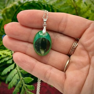 May include: A green oval pendant necklace with a silver chain. The pendant has a dark green center with a lighter green border. The necklace is held in a hand with rings on the fingers. The background is blurred green foliage.