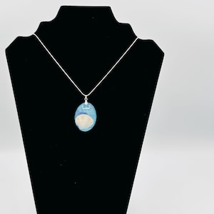 May include: A silver necklace with an oval pendant. The pendant features a light blue and white design, possibly resin, with a seashell element. The necklace is displayed on a black stand against a white background.