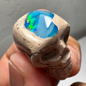May include: A carved skull figurine made of tan stone, with a cavity filled with a vibrant, iridescent opal. The opal displays shades of blue, green, and yellow. The skull has detailed facial features and a textured surface.