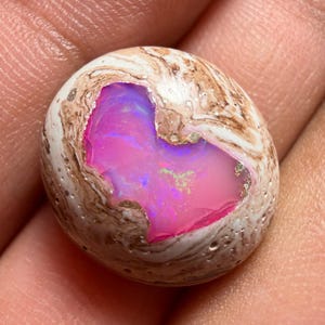 Puede incluir: Una piedra preciosa de ópalo pulida, de forma ovalada. La capa exterior es de color blanco crema con vetas marrones. El centro revela un interior rosa y púrpura vibrante con destellos iridiscentes.