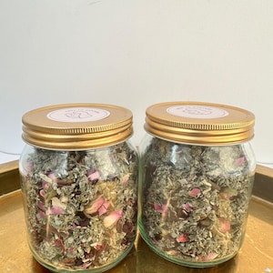 Könnte beinhalten: Zwei klare Gläser gefüllt mit getrockneten Kräutern und Rosenblättern. Die Gläser haben goldfarbene Deckel mit einem rosa Etikett. Die Gläser stehen auf einem goldfarbenen Tablett. Der Inhalt der Gläser ist eine Mischung aus Grün und Rosa.
