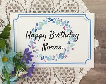 Op de afbeelding: Een witte kaart met een blauwe rand en een aquarelkrans van blauwe, roze en groene bloemen. De tekst "Happy Birthday Nonna" is geschreven in zwarte cursieve letters.