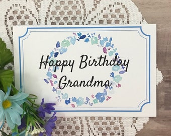 May include: A white card with a blue border and a watercolor floral wreath. The text "Happy Birthday Grandma" is written in black cursive.