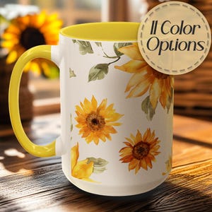 May include: A white ceramic mug with a yellow handle and interior, adorned with watercolor sunflowers and green leaves. The mug has the text "11 Color Options" on a circular sticker. The mug is on a wooden surface.