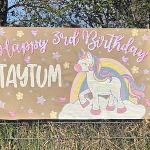 May include: A brown banner with the text "Happy 3rd Birthday TAYTUM" in pink and white. A unicorn with a rainbow mane and tail is on the right. The banner is decorated with hearts, stars, and flowers.