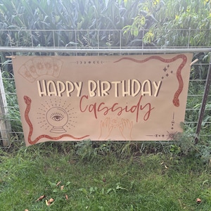 May include: A rectangular tan banner featuring the text "HAPPY BIRTHDAY Cassidy" in dark brown and red script. The design includes illustrations of a crystal ball, tarot cards, snakes, hands, and celestial symbols printed in a dark brown ink.
