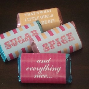 May include: Four wrapped candy bars with text. One bar is yellow with the text "THAT'S WHAT LITTLE GIRLS ARE MADE OF!". Two bars have a light blue and pink striped design with the words "SUGAR" and "SPICE". The last bar is pink with the text "and everything nice..."