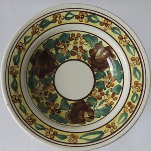 Puede incluir: Un cuenco redondo de cerámica con interior crema y una ancha banda amarilla decorada con flores marrones y hojas verdes. El centro del cuenco es crema, y el borde es blanco con un borde marrón. El cuenco tiene un diseño floral.