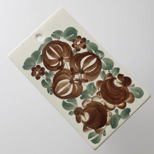 Könnte beinhalten: Rechteckiges Keramik-Schneidebrett mit cremefarbenem Hintergrund. Das Brett hat ein Blumendesign mit braunen und weißen Blumen, grünen Blättern und kleinen braunen Blumenakzenten. Oben befindet sich ein Loch zum Aufhängen.