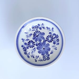 Könnte beinhalten: Eine runde weiße Keramikschale mit einem handgemalten blauen Blumenmuster. Das Design zeigt große zentrale Blüten, umgeben von kleineren Blumen, Blättern und Ranken. Ein breiter blauer Streifen verläuft entlang des inneren Randes der Schale.