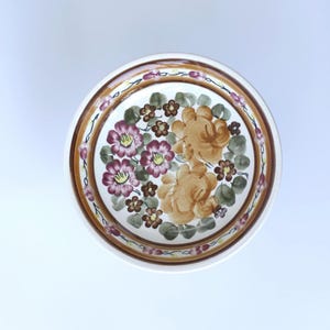 Può includere: Una ciotola rotonda in ceramica con un motivo floreale dipinto a mano, caratterizzato da grandi fiori viola e marrone dorato, fiori più piccoli marroni e foglie verdi. Il bordo è decorato con una fascia marrone e piccoli boccioli viola.