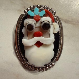 May include: Oval-shaped silver-toned brooch featuring a Santa Claus design. The Santa face is crafted with white, red, black, and turquoise-colored stones. The brooch has a beaded border and a black background.