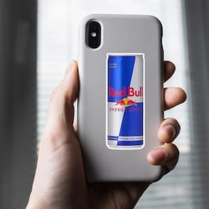 May include: A gray phone case with a Red Bull energy drink sticker. The sticker features a blue and white design with the Red Bull logo and the words "ENERGY DRINK". The phone is held in a person's hand.