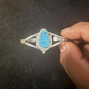 May include: Silver-toned bracelet with a central turquoise-colored inlay, accented with small red and blue details. The bracelet has a teardrop-shaped design and is held by a hand against a dark background. The bracelet's design is intricate and detailed.