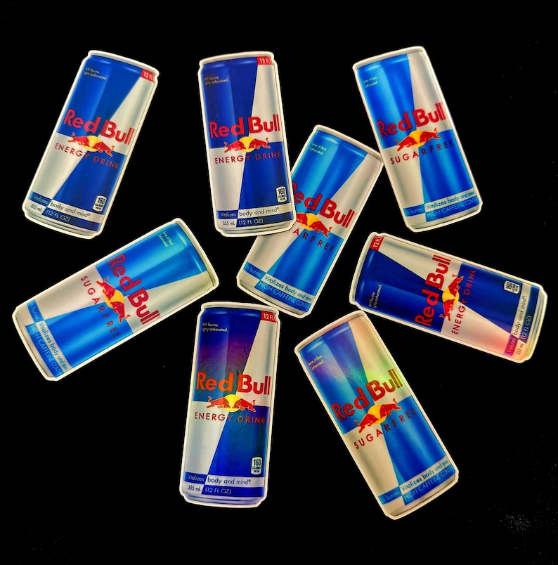 May include: An assortment of Red Bull energy drink cans, featuring a blue and silver design with the Red Bull logo and text. Some cans are labeled "SUGARFREE". The cans are arranged against a black background.