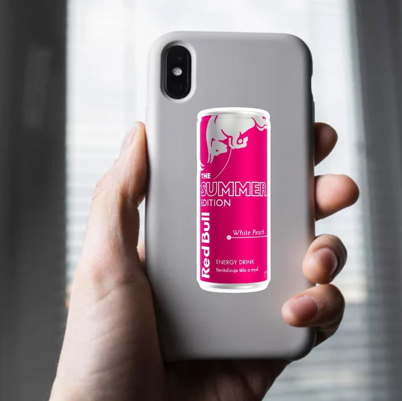 May include: A pink and white Red Bull Summer Edition energy drink can with "White Peach" text, held against a gray phone case. The can features the Red Bull logo and the words "ENERGY DRINK".