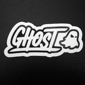 May include: White sticker with the word "GHOST" in a bold, stylized font, accompanied by a small ghost illustration. The sticker has a black outline and is set against a black background.