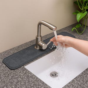 May include: A gray absorbent mat is placed under a brushed nickel faucet. Water flows from the faucet into a white sink. The mat is rectangular and designed to catch water. The sink is set into a speckled countertop.