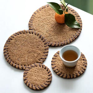 Puede incluir: Un juego de cuatro posavasos redondos de fibras naturales tejidas, con un borde cosido marrón. Un posavasos contiene una taza de té blanca con té, y otro una pequeña naranja con hojas verdes.
