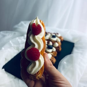 Puede incluir: Primer plano de un eclair de pastelería con glaseado de crema blanca y frambuesas frescas. El eclair se sostiene sobre una superficie negra, con otros eclairs en el fondo. El fondo es una tela blanca drapeada.