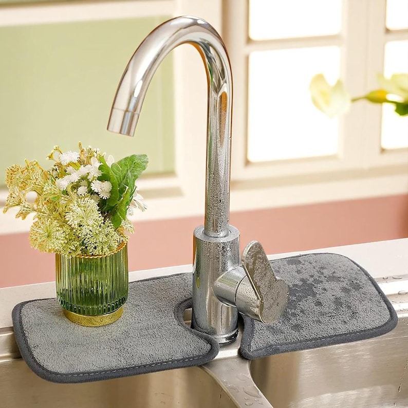 May include: A stainless steel kitchen faucet with a gray absorbent mat designed to catch water. A small vase with flowers sits to the left of the faucet. The mat has a dark gray border and is placed around the faucet base.