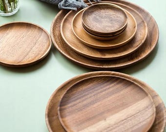 May include: A collection of wooden plates and bowls in various sizes. The plates have a natural wood grain finish, with a warm brown color. The arrangement includes a small stack of bowls and several larger plates, suitable for serving food.